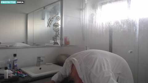 Media: A video shows a person with a towel on their head, leaning over a bathroom sink with toiletries. The bathroom has white tiles, a shower, and a mirror.