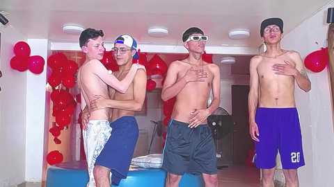 Media: Video of four shirtless young men in swim trunks, three wearing red heart-shaped balloons, embracing in a brightly lit hallway with red balloons on the wall.