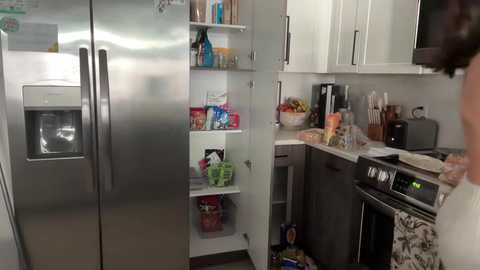 Media: A video juxtaposing a stainless steel fridge and a modern, organized kitchen with white cabinets and black appliances, showcasing a woman in a white dress and apron, preparing food.