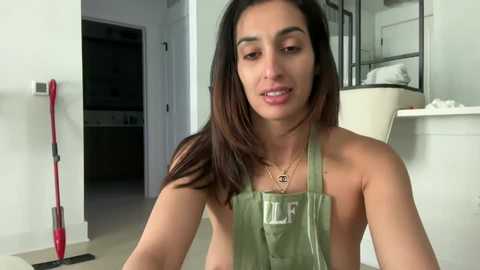 Media: Video of a young, attractive woman with long dark hair, wearing a green apron, in a modern, white kitchen, holding a laundry basket, with a red mop leaning against the wall.