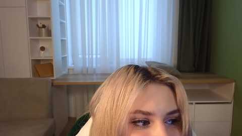 Media: Video of a blonde woman with straight hair, light makeup, and green shirt, gazing out a window with sheer white curtains and a wooden desk.