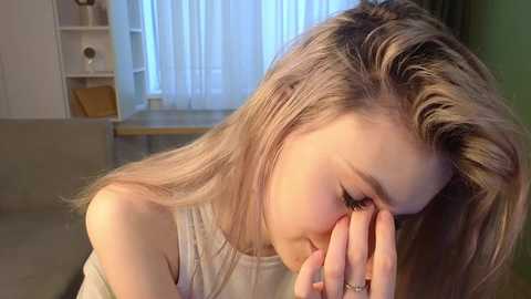 Media: A close-up video of a young woman with light brown hair, wearing a sleeveless top, covering her eyes with her hands, seated in a living room with white curtains and a beige sofa.