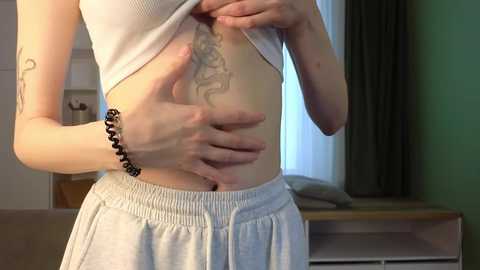 Media: Video of a fair-skinned woman lifting her white crop top, revealing a tattoo on her side. She wears light gray shorts and a black beaded bracelet. Background shows a green wall and wooden dresser.