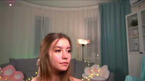 A video of a young girl with light skin and straight brown hair, wearing a light-colored dress, standing in a softly lit, cozy room with pastel-colored balloons, fairy lights, and a white curtain.