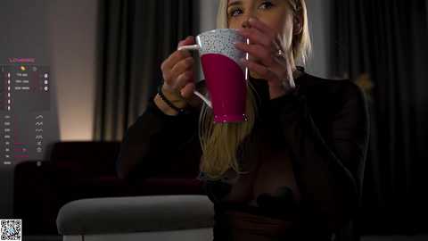 Media: Video of a blonde woman in a sheer black top holding a pink mug, set in a dimly-lit, modern living room with dark curtains and a TV displaying a \"BBC News\" logo.