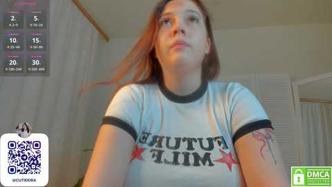 Media: Video of a young woman with fair skin and red hair, wearing a wet, white \"FUCK THIS LIMB\" t-shirt, in a dimly lit room.