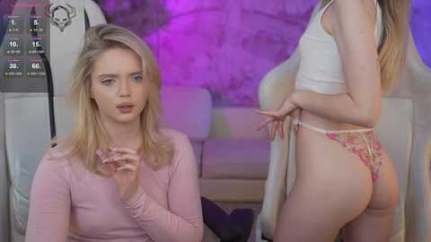 Media: Video of a blonde woman in a pink top, and a brunette woman in floral panties, both seated in a dimly lit room with a purple background.