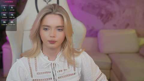 Video of a young Caucasian woman with straight blonde hair, wearing a white blouse with intricate embroidery, sitting indoors. Background features a purple wall and a beige couch.