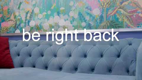 Media: Video of a modern bedroom with a tufted, light blue velvet headboard, bright floral wallpaper, and a \"be right back\" text overlay.