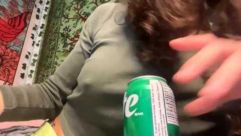 Media: Video of a woman with curly brown hair, wearing a green long-sleeve top, holding a green Coke can in her right hand, in a colorful, tapestry-decorated room.