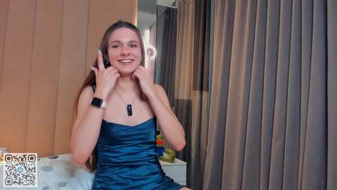 Media: Video of a smiling young woman with long brown hair, wearing a sleeveless blue satin dress, holding a smartphone to her ear. Background features beige wall, grey curtains, and a white sofa.