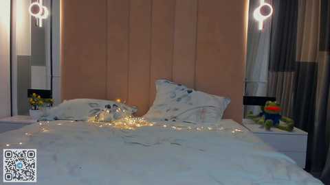 Media: Video of a modern bedroom with a white bed, white duvet, and decorative pillows. The headboard is a light wood panel with two hanging circular lights. Nightstands hold yellow flowers and a colorful toy frog.