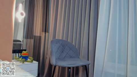 Media: Video of a modern bedroom with a gray, quilted chair, beige curtains, a white nightstand, and a colorful toy on top.