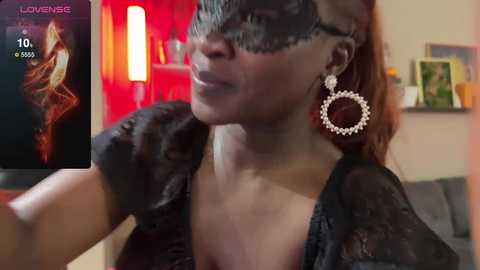 Media: Video of a dark-skinned woman with a mysterious black mask, adorned with large, ornate earrings, posing seductively indoors. The background features a red wall and a blurred bookshelf.