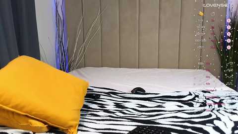 Media: Video of a modern bedroom with a zebra-print bedspread, yellow pillow, beige padded headboard, and decorative green plants on the nightstand.