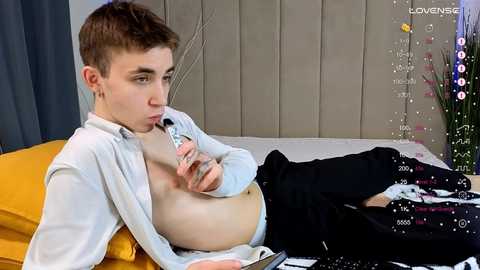 Media: Video of a shirtless, fair-skinned teenage boy with short brown hair, wearing black pants, lying on a bed with a mustard-yellow pillow. He is using a sex toy. Background includes a gray curtain, a white radiator, and a colorful vase.