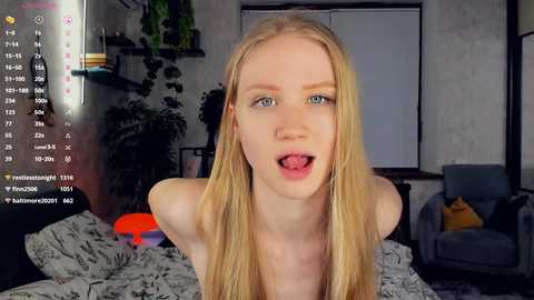 Media: Video of a pale, blonde, nude young woman with blue eyes, sticking out her tongue, in a modern bedroom with grey bedding, a green plant, and a gray armchair.