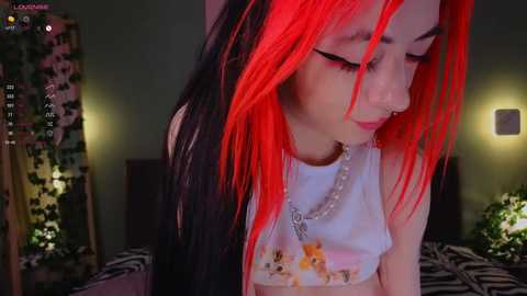 Media: Video of a young woman with long, black and bright red hair, wearing a white tank top with cartoon characters, in a dimly lit room.