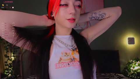 Media: Video of a young, pale-skinned woman with long, dyed red and black hair, wearing a \"Sailor Moon\" T-shirt, silver necklace, and black jacket, posing in a dimly lit bedroom with green walls and a bed.