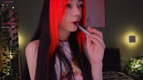 Media: Video of a pale-skinned, slim Asian woman with long, straight black hair streaked with bright red, wearing a white graphic tee and black choker, seductively licking a lipstick applicator, in a dimly lit bedroom with a zebra print bedspread.