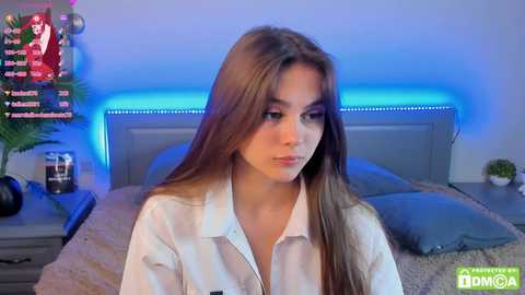 Media: Video of a young woman with long brown hair, wearing a white shirt, seated in a bedroom with blue LED lighting and a bed with gray bedding.