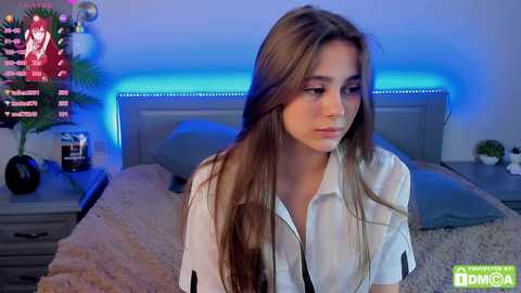 Media: Video of a young woman with long brown hair, wearing a white shirt, seated on a bed with a beige blanket. Background features a TV displaying a red screen, and a wooden nightstand with a plant.