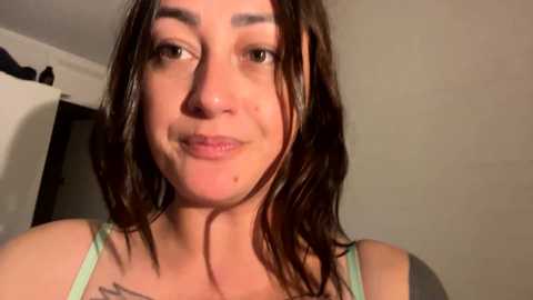 Media: A close-up video of a young woman with wet, shoulder-length brown hair, fair skin, and a slight smile. She wears a green tank top. The background is a plain beige wall.