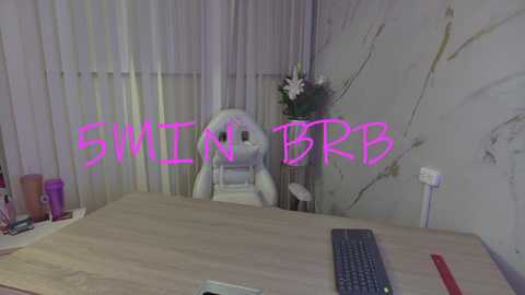 Media: Video of a white rabbit on a desk chair, with a computer keyboard and colorful office supplies, in a room with vertical blinds and a floral wall. Text \"SMILE & BRRR\" overlaid.