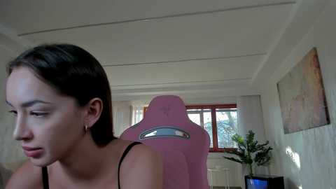 Media: A video shows a young woman with fair skin, straight brown hair, and a slender physique, wearing a black spaghetti strap top, seated in a pink gaming chair in a brightly lit room with white walls and a window.