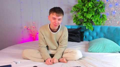 Media: Video of a young man with short brown hair, wearing a beige sweater and light pants, sitting cross-legged on a bed with turquoise headboard and green pillow, surrounded by string lights and a green plant.