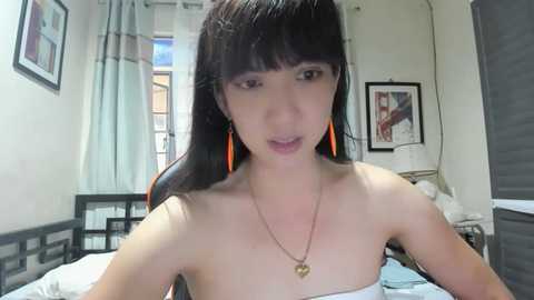 Media: Video of an Asian woman with long black hair, wearing orange earrings, a white strapless top, and a gold heart necklace, standing in a bedroom with framed art and a bed.