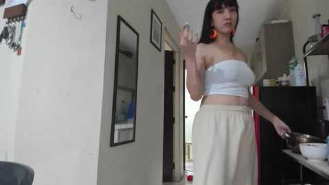 Media: Video of an East Asian woman with long black hair and fair skin, wearing a white strapless top and beige high-waisted pants, gesturing in a modern kitchen with white walls and black appliances.