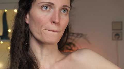 Media: Video of a fair-skinned woman with blue eyes and long dark hair, smiling softly. She has a delicate, subtle makeup look. The background features a blurred, cozy room with warm lighting and a decorative mirror.