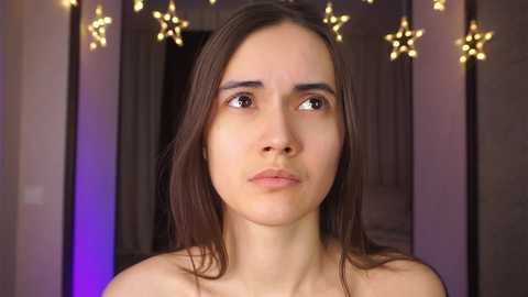 Media: Video of a young woman with light skin and straight, shoulder-length brown hair, wearing a neutral expression. She is topless, with a blurred background featuring star-shaped fairy lights.