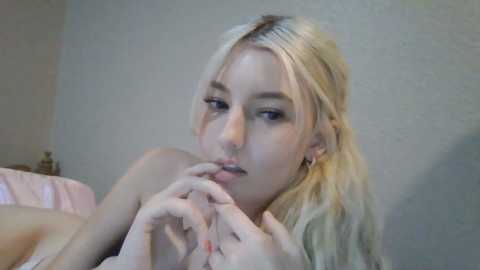 Media: Video of a topless, blonde woman with fair skin, light makeup, and closed eyes, licking her fingers, in a soft-focus bedroom setting with pastel colors.