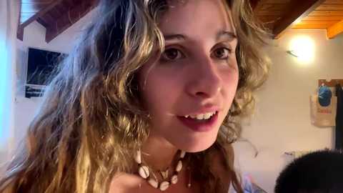 Media: Video of a young woman with wavy, shoulder-length brown hair, wearing a shell necklace, smiling. She stands in a rustic, wooden-beamed room with a warm light.