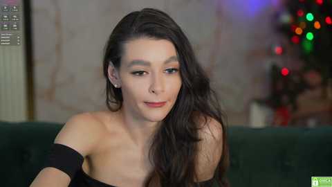 Media: Video of a young, slender woman with long, dark hair, fair skin, and light makeup, wearing a black off-shoulder top, sitting on a dark couch. Christmas lights and a festive background in the blurred background.