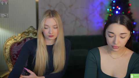 Media: Video of two young women; one blonde with long hair, the other brunette with dark hair, sitting on a green couch, looking at a smartphone, in a cozy, festive room with a Christmas tree.