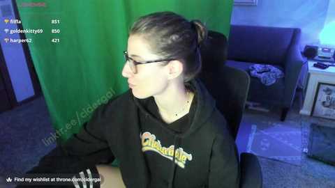 Media: Video of a young woman with glasses, light skin, and brown hair tied back, wearing a black hoodie with a yellow logo, sitting in a dark chair in a dimly lit room with a green screen in the background.