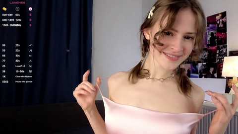 A video of a young, light-skinned woman with brown pigtails, wearing a pink satin top, smiling in a cozy room with a dark curtain, calendar, and posters.