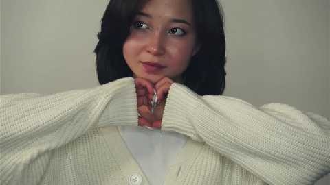 Media: Video of a young woman with fair skin and shoulder-length dark hair, wearing a cream-colored knitted cardigan over a white shirt. She looks perplexed, holding the cardigan open with her hands. The background is plain and neutral.