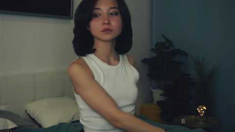 Media: Video of a young woman with short dark hair, wearing a white tank top, sitting on a bed with a beige headboard, in a dimly lit room with a potted plant and a yellow lamp.