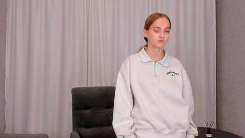 Media: Video of a young Caucasian woman with light skin and brown hair tied back, wearing a white polo shirt and seated in a black office chair against gray curtains and a concrete wall.