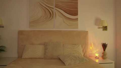 Media: Video of a minimalist bedroom with beige walls, a light beige headboard, abstract wave-patterned wall art, two matching wall sconces, and a lit candle on a nightstand.