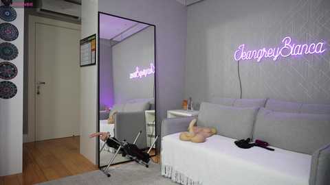 Media: A video of a chic, minimalist bedroom with a large mirror, neon \"EvepyoBianca\" sign, white bedding, and a small chair with a stroller beside it.