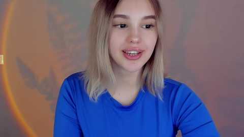 Media: Video of a young woman with fair skin and shoulder-length blonde hair, smiling, wearing a blue long-sleeved shirt. Background is blurred with an orange, circular light source.