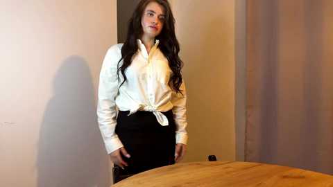 Media: Video of a young woman with long brown hair, wearing a white button-up shirt tied at the waist and black pants, standing in a minimalist room with beige walls and a wooden table.