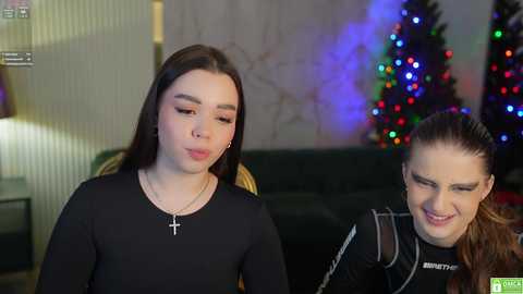 Media: Video of two young women, one with long brown hair in a black dress, the other with auburn hair, smiling, in a cozy living room with a Christmas tree and colorful lights.