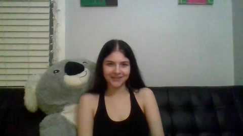 Media: A video of a young woman with long black hair, wearing a black tank top, sitting on a black leather couch. She smiles while a large, gray, plush kangaroo rests on her left shoulder.