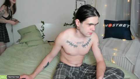 Media: Video of a shirtless, pale-skinned man with long dark hair, sitting on a bed, wearing gray plaid pajama pants. He has a cannabis leaf tattoo on his chest. A woman in a black dress stands in the background.